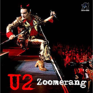 May include: A performer in a gold metallic suit and boots, holding a microphone and a stand, with the band name "U2" and the word "Zoomerang" in red and white text. A crowd is visible in the background.