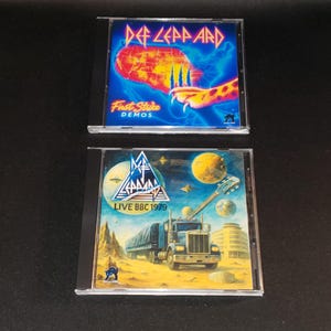 May include: Two Def Leppard CD albums. The top CD is titled "First Strike Demos" with a colorful design. The bottom CD is "Live BBC 1979" with a truck and guitar graphic. Both CDs are in clear plastic cases.