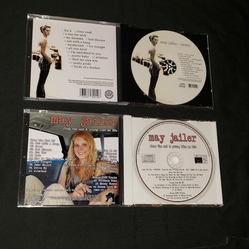 May include: Four CD cases featuring the music of May Jailer. The front cover of the CD case on the left features a woman with blonde hair and a black shirt. The text on the CD case reads "May Jailer from the end & young like me EP". The front cover of the CD case on the right features a woman with blonde hair and a black shirt. The text on the CD case reads "May Jailer from the end & young like me EP". The front cover of the CD case in the middle features a woman with blonde hair and a black shirt. The text on the CD case reads "May Jailer - Sirens".