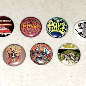 May include: Seven round, colorful, pinback buttons with the band name "Gov't Mule" printed on each. The buttons feature various designs, including a car, a sun, a musical instrument, and a sheep.