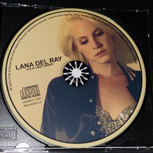 Puede incluir: Un CD con una mujer de cabello rubio que lleva una chaqueta negra y un top brillante. El texto en el CD dice "LANA DEL RAY A.K.A. LIZZY GRANT" y "COMPACT DISC DIGITAL AUDIO Made in USA REMASTER 2.0".