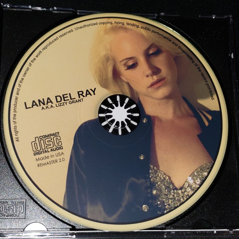 Puede incluir: Un CD con una mujer de cabello rubio que lleva una chaqueta negra y un top brillante. El texto en el CD dice "LANA DEL RAY A.K.A. LIZZY GRANT" y "COMPACT DISC DIGITAL AUDIO Made in USA REMASTER 2.0".