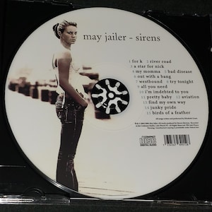 Puede incluir: Un CD blanco con un centro negro y una foto de una mujer con cabello oscuro y una camisa blanca. El texto "may jailer - sirens" est&aacute; impreso en el CD. La lista de canciones est&aacute; impresa debajo del t&iacute;tulo.