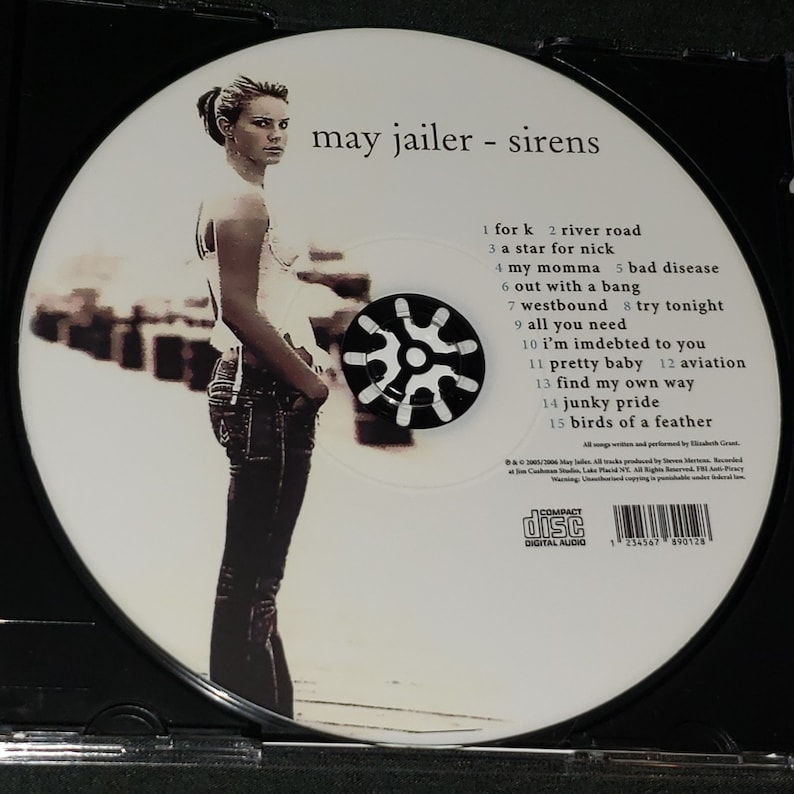 Puede incluir: Un CD blanco con un centro negro y una foto de una mujer con cabello oscuro y una camisa blanca. El texto "may jailer - sirens" est&aacute; impreso en el CD. La lista de canciones est&aacute; impresa debajo del t&iacute;tulo.