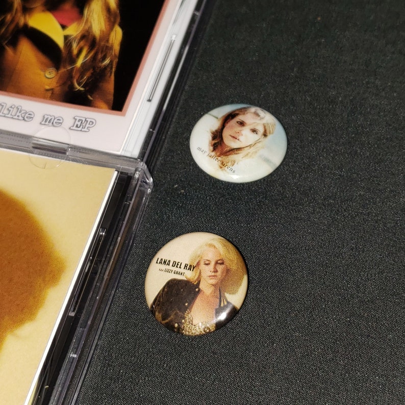 May include: Two round button pins featuring images of singers Lana Del Rey and May Jailer. The Lana Del Rey pin has the text "Lana Del Rey" and "Ultraviolence" on it. The May Jailer pin has the text "May Jailer" on it.