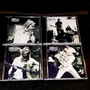 May include: Four Nirvana CD albums. Each CD case features black and white images of the band members. The album titles include "Home Demos", "Studio Sessions", "Broadcasts", and "Live Rarities". The Nirvana logo is displayed on each CD case.