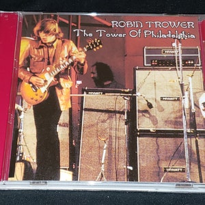 May include: A red CD case with a picture of a man playing a guitar on stage. The text on the case reads "Robin Trower The Tower of Philadelphia".
