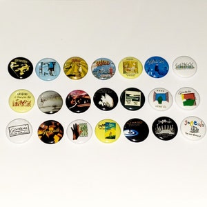 May include: A collection of 18 round buttons featuring various album covers from the band Genesis. The buttons are colorful and feature images of the band members, album titles, and song lyrics.
