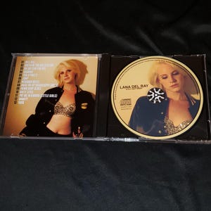 May include: A CD case with a photo of a woman wearing a blue jacket and a sparkly bra. The CD is labeled "Lana Del Ray" and has a black and white sunburst design. The tracklist on the back of the case includes songs like "Kill Kill", "Queen of the Gas Station", and "Yayo".