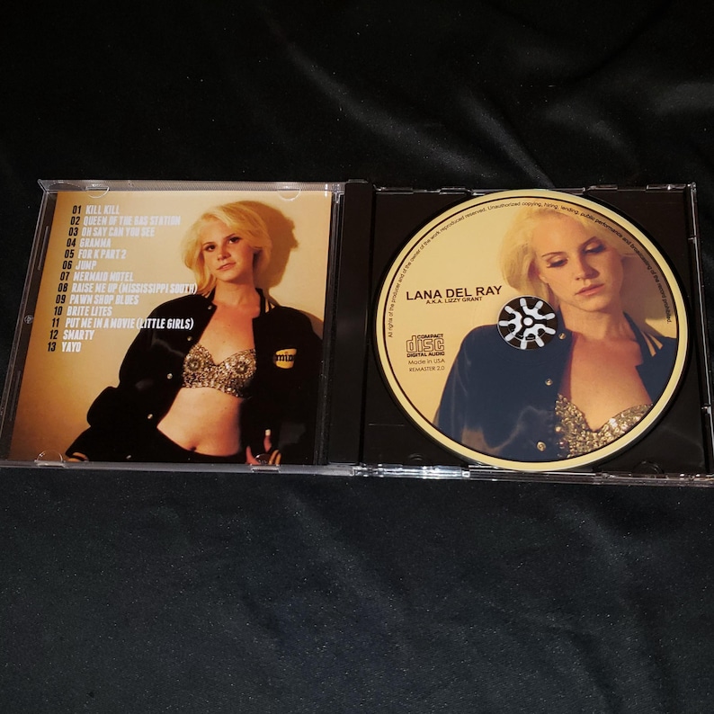 May include: A CD case with a photo of a woman wearing a blue jacket and a sparkly bra. The CD is labeled "Lana Del Ray" and has a black and white sunburst design. The tracklist on the back of the case includes songs like "Kill Kill", "Queen of the Gas Station", and "Yayo".