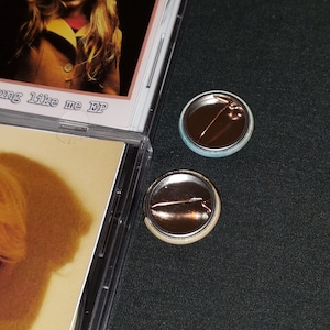 May include: Two back-to-back pinback buttons with a silver metal backing and a pin. The button on the left has a light blue background and the button on the right has a light beige background.