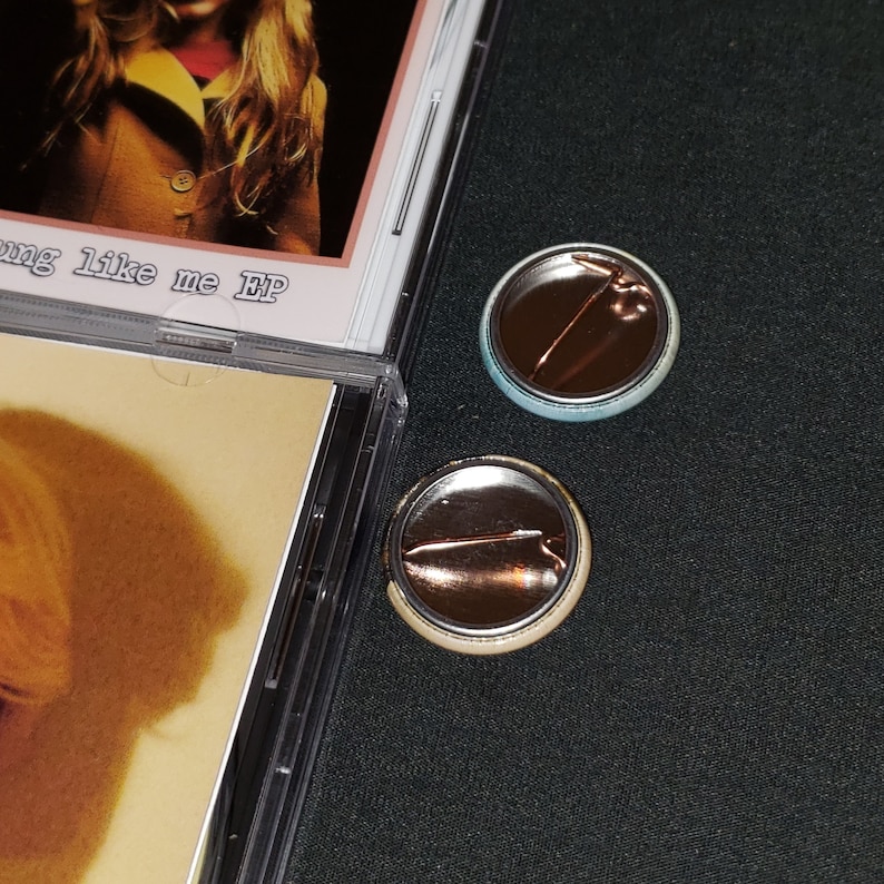 May include: Two back-to-back pinback buttons with a silver metal backing and a pin. The button on the left has a light blue background and the button on the right has a light beige background.