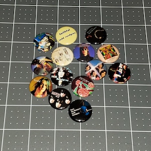 May include: A collection of 13 round buttons featuring album covers by musician Todd Rundgren. The buttons feature various album titles including "Faithful", "Healing", "Runt", "The Ballad of Todd Rundgren", "Todd", "Todd Rundgren", and "A Wizard, A True Star".