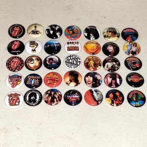 May include: A collection of 25 round button pins featuring images of the Rolling Stones band members and album covers. The pins are colorful and feature various images, including the band's iconic tongue logo, band members' portraits, and album covers.