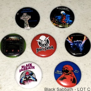 May include: A collection of seven Black Sabbath band pinback buttons. The buttons feature different album covers and logos. The buttons are round and have a black background. The buttons are arranged in a circle on a white surface.