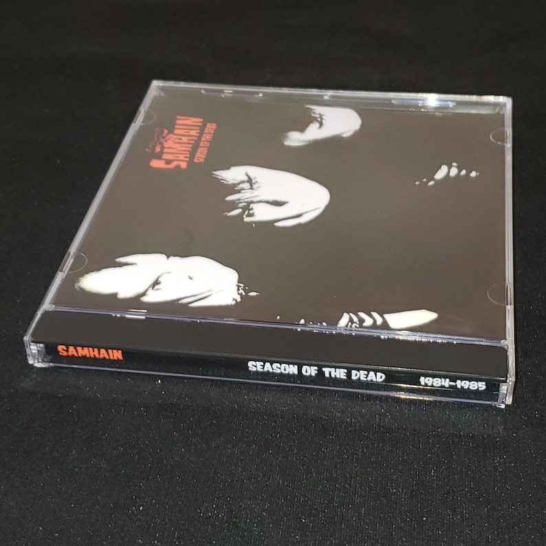 May include: A black CD case with a black and white image of a band on the front. The title "SAMHAIN" is printed in red on the front of the case. The title "SEASON OF THE DEAD 1984-1985" is printed in white on the spine of the case.