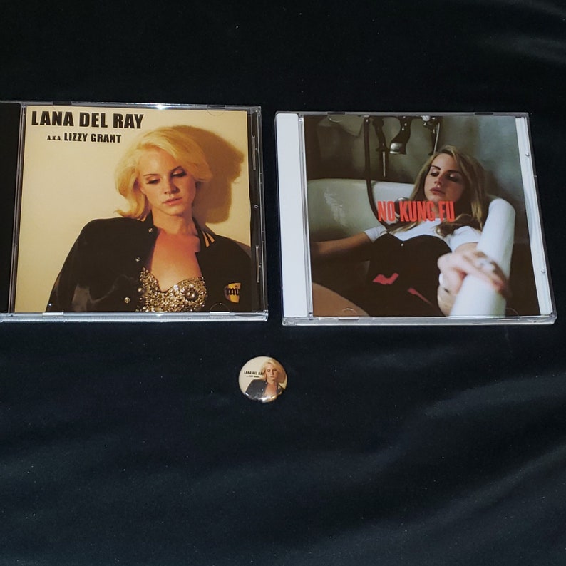 Puede incluir: Dos discos compactos con portadas de &aacute;lbum que muestran a una mujer con el pelo corto y rubio. La primera portada del &aacute;lbum se titula "Lana Del Ray - AAA Lizzy Grant". La segunda portada del &aacute;lbum se titula "No Kung Fu". Un peque&ntilde;o bot&oacute;n con una imagen de la mujer est&aacute; en el centro de la imagen.