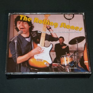 May include: CD case with a photo of The Rolling Stones band members. The text on the case reads "The Rolling Stones The Complete Woodstock Rehearsal Tapes".