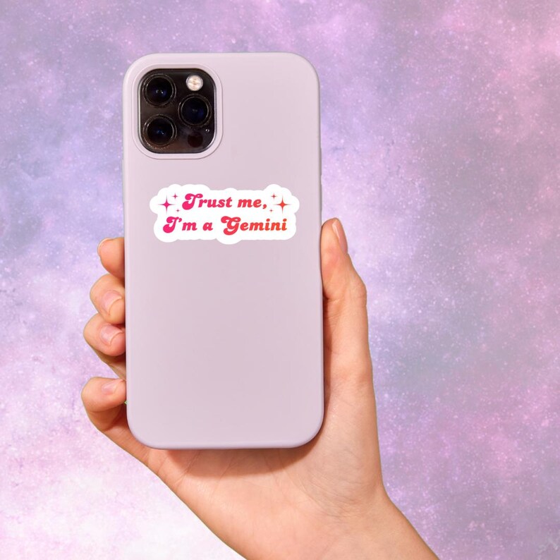 May include: A light purple phone case with a sticker that reads "Trust me, I'm a Gemini" in pink text. The sticker has a white border and star accents. The phone case is held in a hand against a purple and pink background.