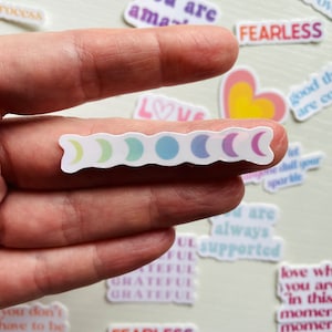 May include: A sticker sheet with a row of six crescent moon stickers in various pastel colors. The stickers are on a white background and are being held by a hand.