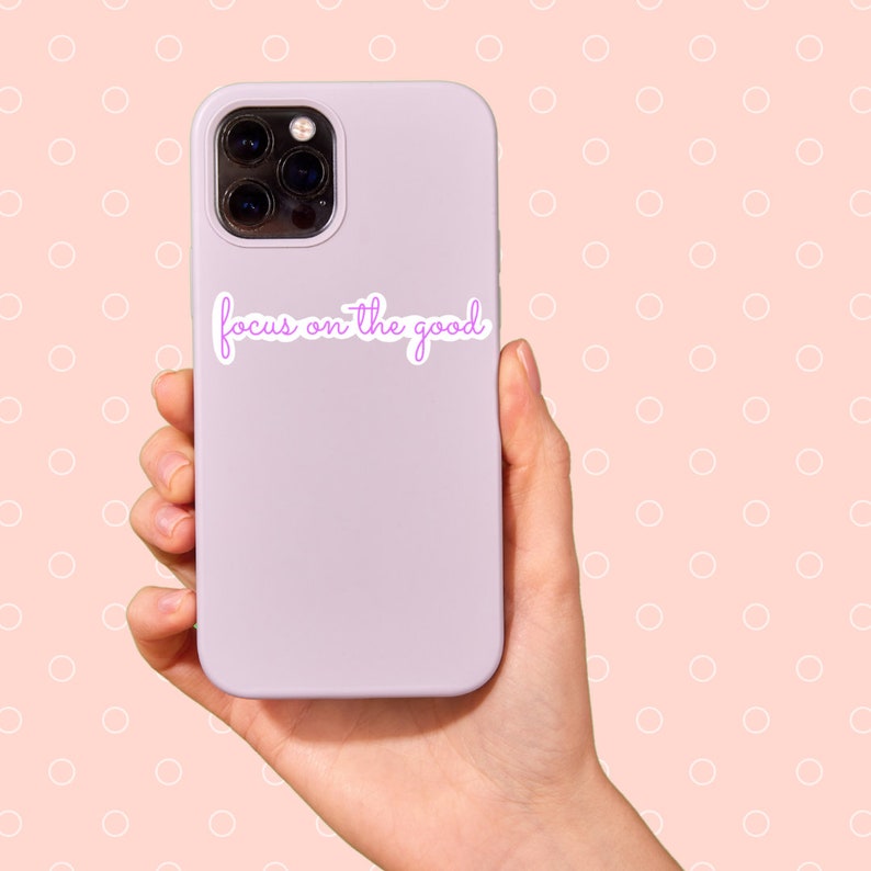 May include: A lilac phone case with a sticker reading "focus on the good" in cursive script. The phone, with a multi-lens camera, is held in a hand against a peach backdrop with white polka dots.