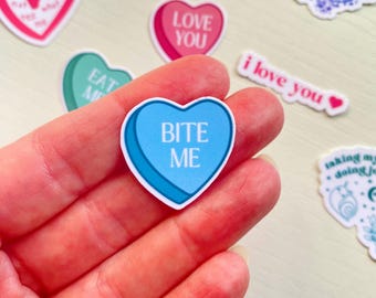 Może przedstawiać: Niebieska naklejka w kształcie serca z napisem "BITE ME" białymi literami. Naklejka jest trzymana w dłoni.