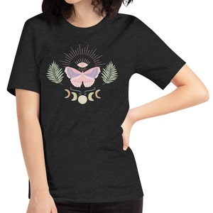 Puede incluir: Una camiseta gris oscuro jaspeada con un dise&ntilde;o de mariposa rosa y morada. La mariposa tiene un ojo en el centro de sus alas. El dise&ntilde;o tambi&eacute;n incluye una luna creciente y un sol con rayos.
