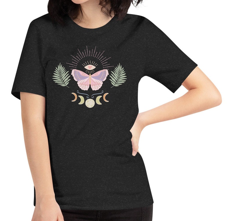 Puede incluir: Una camiseta gris oscuro jaspeada con un dise&ntilde;o de mariposa rosa y morada. La mariposa tiene un ojo en el centro de sus alas. El dise&ntilde;o tambi&eacute;n incluye una luna creciente y un sol con rayos.