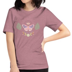 Puede incluir: Una camiseta malva con un dise&ntilde;o de mariposa rosa. La mariposa tiene un ojo en el centro de sus alas y est&aacute; rodeada de un sol radiante, lunas crecientes y hojas de palma.