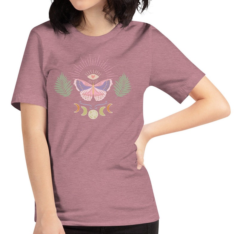 Puede incluir: Una camiseta malva con un dise&ntilde;o de mariposa rosa. La mariposa tiene un ojo en el centro de sus alas y est&aacute; rodeada de un sol radiante, lunas crecientes y hojas de palma.