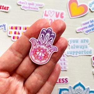 May include: A sticker with a hamsa hand design. The hamsa is white with a pink and purple ombre effect. The hamsa has a detailed design with a large eye in the center. The sticker is on a hand.