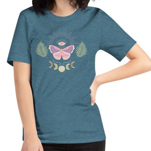 Puede incluir: Una camiseta azul turquesa con un dise&ntilde;o de mariposa rosa. La mariposa tiene un tercer ojo y un coraz&oacute;n en sus alas. El dise&ntilde;o tambi&eacute;n incluye una luna creciente y una luna llena.