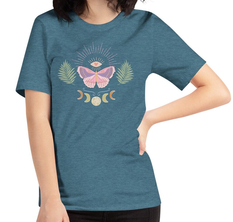 Puede incluir: Una camiseta azul turquesa con un dise&ntilde;o de mariposa rosa. La mariposa tiene un tercer ojo y un coraz&oacute;n en sus alas. El dise&ntilde;o tambi&eacute;n incluye una luna creciente y una luna llena.