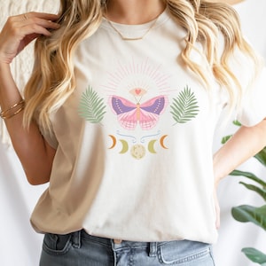 Puede incluir: Una camiseta blanca con una mariposa rosa, lunas crecientes y hojas de palma impresas en la parte delantera. La mariposa tiene un dise&ntilde;o morado y azul con un coraz&oacute;n rosa en el centro.