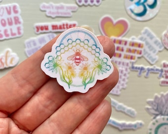 May include: A rainbow-colored sticker with a bee and flowers inside a honeycomb design. The sticker is held in a hand.