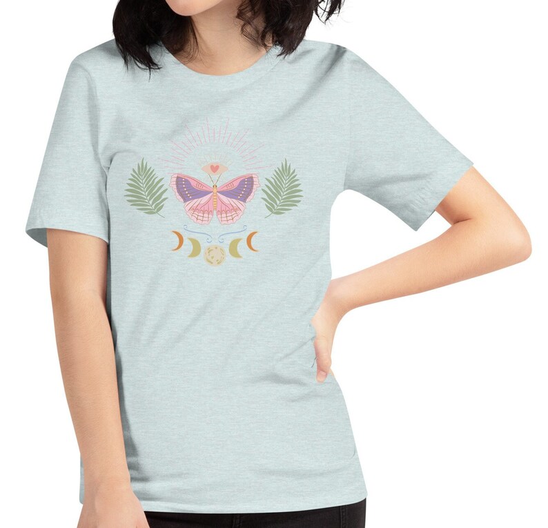 Puede incluir: Una camiseta azul claro con un gr&aacute;fico de mariposa rosa, rodeada de hojas y lunas crecientes. La mariposa tiene un coraz&oacute;n en el centro de sus alas.