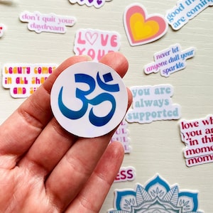 May include: A white sticker with a blue and teal Om symbol. The sticker is being held by a hand. The sticker is surrounded by other stickers with positive messages.