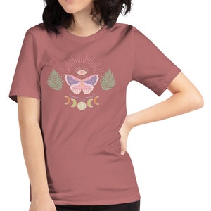 Puede incluir: Una camiseta de manga corta de color malva con un dise&ntilde;o gr&aacute;fico de una mariposa con un tercer ojo y un coraz&oacute;n, rodeada de lunas crecientes y hojas de palma.