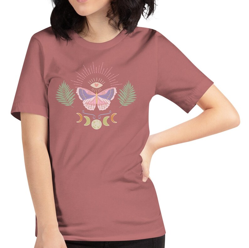 Puede incluir: Una camiseta de manga corta de color malva con un dise&ntilde;o gr&aacute;fico de una mariposa con un tercer ojo y un coraz&oacute;n, rodeada de lunas crecientes y hojas de palma.