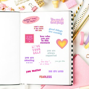 May include: An open notebook with motivational quote stickers in pinks, purples, and yellows. The notebook is surrounded by stationery items such as pens, washi tape, and paperclips, creating a colourful, organised scene.