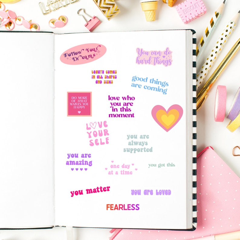 May include: An open notebook with motivational quote stickers in pinks, purples, and yellows. The notebook is surrounded by stationery items such as pens, washi tape, and paperclips, creating a colourful, organised scene.