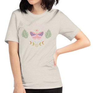Puede incluir: Una camiseta gris claro con una mariposa rosa, hojas verdes y un dise&ntilde;o de luna creciente. La mariposa tiene alas moradas y un peque&ntilde;o coraz&oacute;n en el centro.