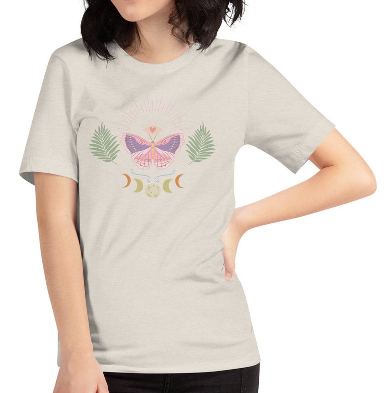 Puede incluir: Una camiseta gris claro con una mariposa rosa, hojas verdes y un dise&ntilde;o de luna creciente. La mariposa tiene alas moradas y un peque&ntilde;o coraz&oacute;n en el centro.