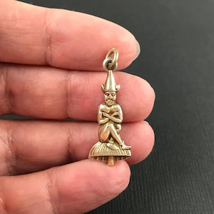 May include: A metallic gold-coloured charm pendant featuring a seated figure with a pointed hat and crossed arms, perched atop a textured mushroom-shaped base. The charm includes a small circular jump ring at the top for attachment to a chain.