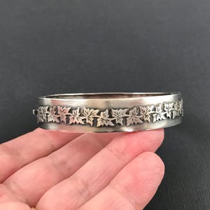 May include: Silver hinged bangle bracelet featuring a repeating leaf design. The bracelet is displayed on a hand against a dark backdrop. The leaves are detailed, forming a decorative border around the bracelet.