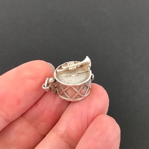May include: A small, silver charm depicting a drum with a hinged lid and a trumpet. The drum has a lattice pattern around the sides. The trumpet is attached to the lid. The charm is held in a person's hand.