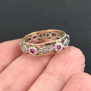 May include: A two-tone ring featuring a gold band and a silver-toned outer layer with an intricate lattice design. The outer layer is set with small, round, vibrant pink rubies and tiny clear stones arranged in a repeating floral-inspired pattern.