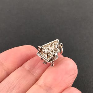 May include: A miniature silver-toned charm shaped like a grand piano, featuring intricate filigree cutouts on the lid, a keyboard section, and small legs. The charm includes a small circular jump ring attached to the back for hanging purposes.