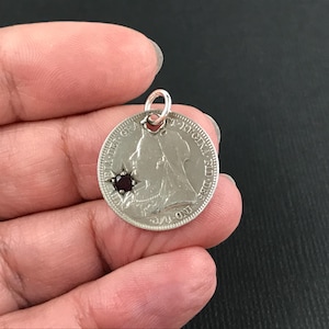 May include: A circular silver-toned coin pendant featuring a profile portrait of a monarch and the text VICTORIA DEI GRA BRITT REGINA FID DEF IND IMP. A small, dark red, round gemstone is set into the left side of the coin, held by a silver jump ring.