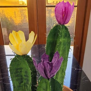 Op de afbeelding: Een cactusplant in een pot met drie kleurrijke papieren bloemen. De bloemen zijn geel, paars en roze. De cactus staat in een terracotta pot met een schotel.